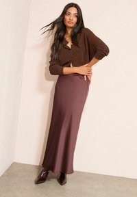 Brązowy teksturowany kardigan z guzikami, zestawiony z długą, gładką, ciemnobordową spódnicą oraz brązowymi, szpiczastymi botkami za kostkę. Minimalistyczne otoczenie.