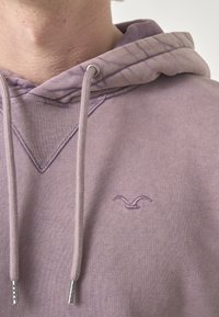 Lila Kapuzenpullover mit Kordelzug und silbernen Metall-Enden. Verfügt über eine V-förmige Naht und ein gesticktes Logo auf der Brust. Weiche Textur.