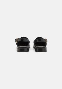 Black patent leather sandals with a low heel, ankle strap, and gold hardware detail. Open back design, featuring a smooth shiny texture.