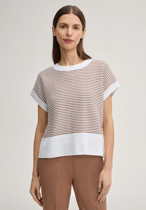 Donna con capelli castano lisci indossa un top a maniche corte beige a righe bianche in maglia e pantaloni marrone chiaro, in piedi davanti a uno sfondo neutro.