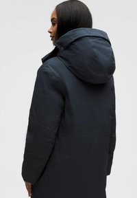 Cappotto imbottito blu navy con cappuccio, caratterizzato da una texture liscia e un design leggermente gonfiato. Include chiusure a pressione e dettagli di cucitura minimi.