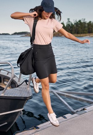 Vrouw stapt van een boot af op een steiger bij het water, draagt een roze t-shirt, zwarte rok, witte sneakers, zwarte pet en draagt een zwarte tas.