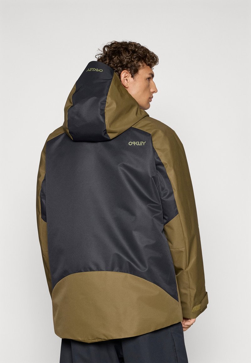 Veste Oakley avec un schéma de couleurs noir et vert olive, capuche et matériau texturé avec une finition lisse. Détail de logo notable au dos.