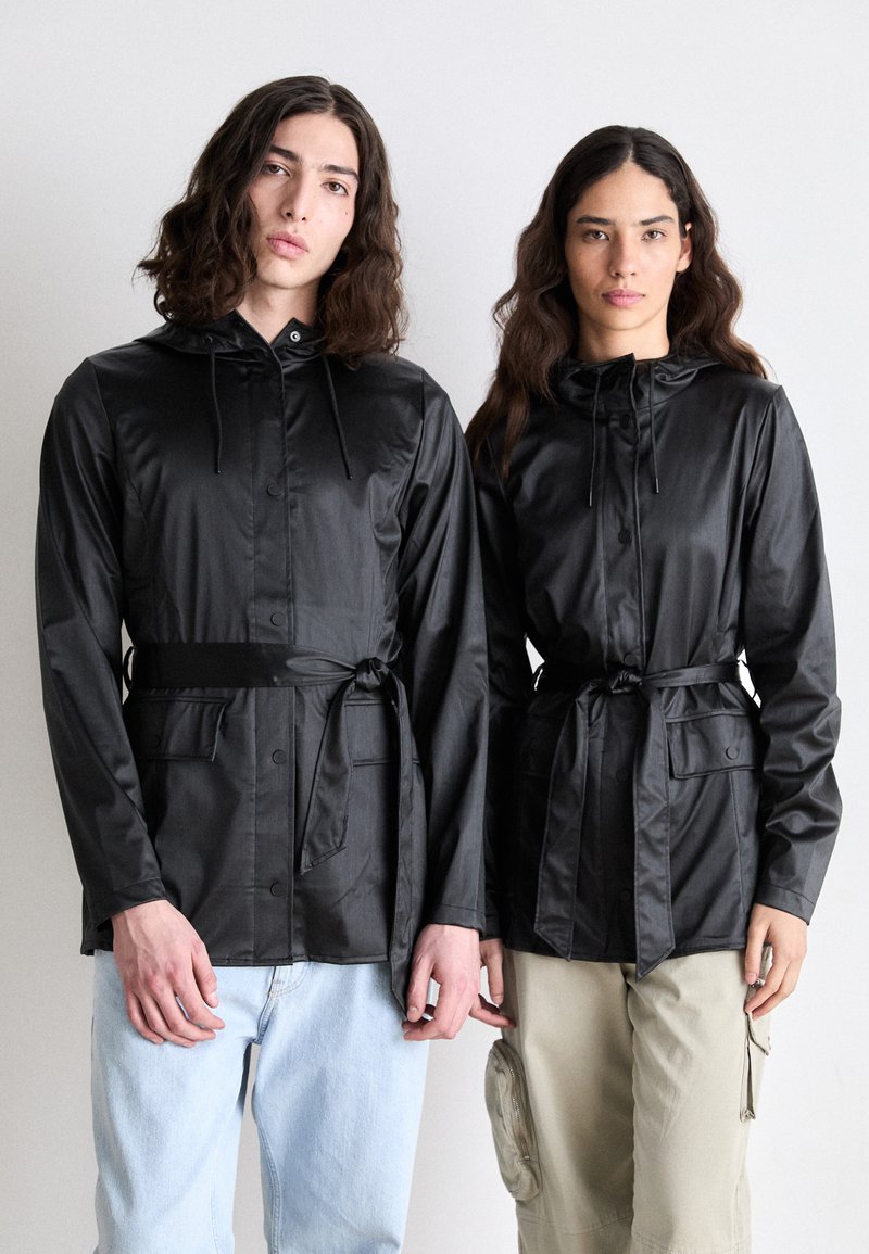 Des vestes légères noires avec capuche, dotées de boutons et de liens à la taille, ainsi que de poches avant. Les mannequins posent dans des attitudes décontractées.