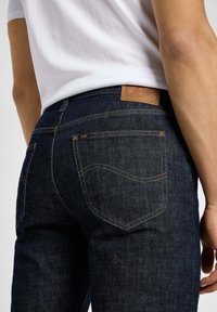 Jeans en denim bleu foncé avec une texture décolorée, des coutures contrastantes et une patch en cuir au niveau de la taille ; présente deux poches arrière.