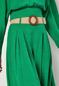 Groene geplooide broek van een gestructureerde stof, voorzien van een geweven beige riem en een houten ronde gesp.