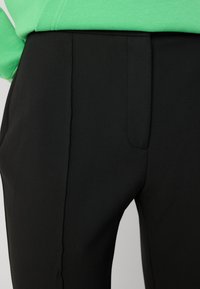 Pantaloni negri ajustați, cu o textură netedă, dotati cu un buzunar lateral și detalii subtile de pliuri. Asortați cu un top verde.