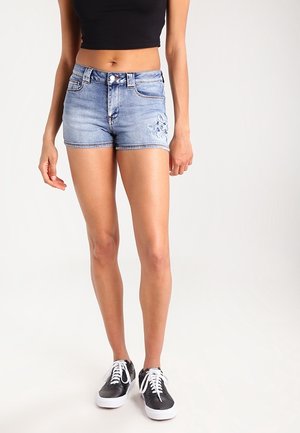 Jeansshorts med en ljusblå tvätt, fransade fållar och blommönsterbroderi på vänster ben, kombinerade med svarta sneakers och en cropped topp.