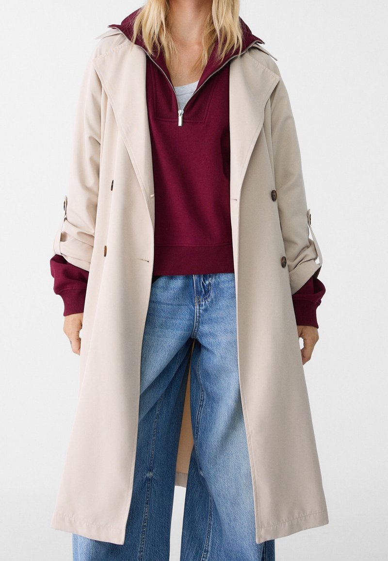 Trench-coat beige avec détails de ceinture, superposé à un sweat à capuche bordeaux et un jean large bleu clair. Texture lisse, boutons visibles en accent.