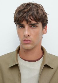 Giovane uomo con capelli corti e mossi di colore castano, occhi verdi, che indossa una giacca beige e un maglione bianco a collo rotondo, espressione neutra, sfondo bianco uniforme.
