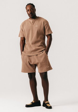 Mann med flettehår iført matchende brun kortermet skjorte og shorts med svarte og beige loafers, stående med hendene i lommene.