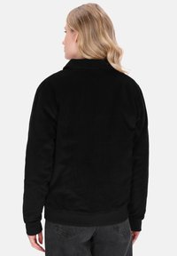 Kvinde med blond hår, der vender væk, iført en sort fløjljakke og mørkegrå jeans foran en simpel lys baggrund.