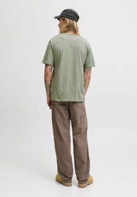 Lys grøn bomuld t-shirt, rund hals, korte ærmer, parret med brune cargo bukser og gule støvler, synlige tatoveringer på armene.