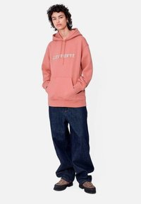 Sudadera rosa con bolsillo frontal y capucha con cordón, combinada con jeans oscuros holgados y zapatos marrón claro. Diseño simple, sin patrones.