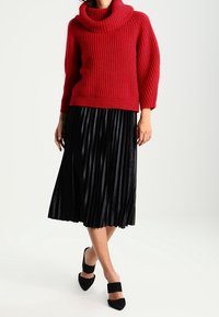 Rode geribde gebreide trui met een colkraag, gecombineerd met een zwarte plissé midi-rok en zwarte chunky hakken.