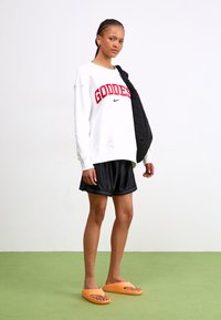 Valkoinen huppari, jossa on punainen "GODDESS" -teksti ja musta Nike-logo, yhdistetty mustiin shortseihin ja oransseihin sandaaleihin. Musta toppatakki roikkuu olkapäällä.