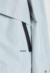 Light blue waterproof jacket featuring a black zip pocket, layered design, and a branded label in black at the bottom. Smooth texture.