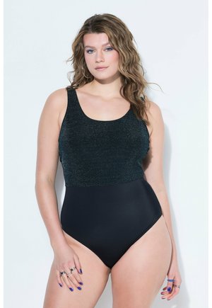 Donna con capelli biondi ondulati che indossa un costume da bagno intero nero senza maniche con una parte superiore scintillante su uno sfondo chiaro.