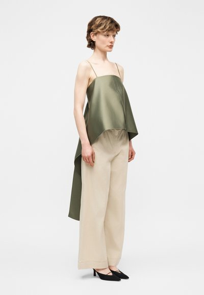 Γυναίκα που φορεί ένα ασύμμετρο τοπ από σατέν σε ελαιί απόχρωση με λεπτές τιράντες, μπεζ παντελόνι με φαρδιά μπατζάκια και μαύρες μυτερές γόβες, στέκεται πλάγια.