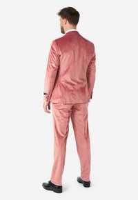 Traje de terciopelo rosa con chaqueta de un solo botón y pantalones a juego. La chaqueta tiene acentos blancos en los puños. Se lleva con zapatos negros.