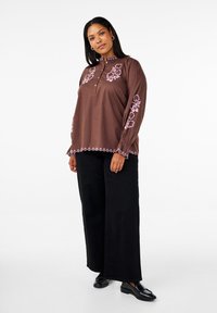 Camicia a maniche lunghe marrone con ricami floreali rosa sulle maniche e sull'orlo, dotata di colletto alto e chiusura con bottoni. Pantaloni neri a gamba larga.