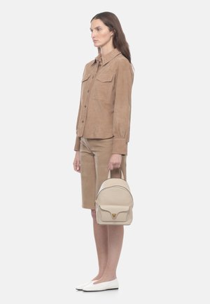 Femme en veste beige boutonnée et short, tenant un petit sac à dos crème, portant des ballerines blanches, debout devant un fond uni.