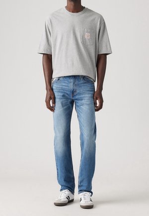 Homme portant un t-shirt gris clair à manches courtes, un jean bleu et des baskets blanches avec des rayures noires, debout devant un fond uni.