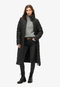 Lang, sort quilted jakke med høj krave, parret med en grå sweater og sorte jeans. Afsluttet med sorte snørebåndsstøvler til anklerne.