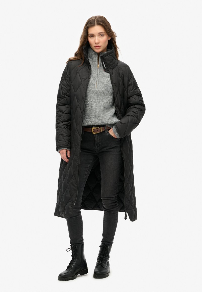 Lang, sort quilted jakke med høj krave, parret med en grå sweater og sorte jeans. Afsluttet med sorte snørebåndsstøvler til anklerne.