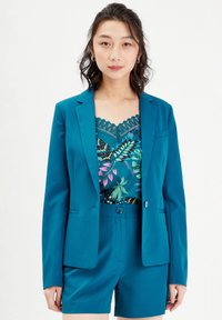 Blazer en sarcelle, tissu lisse, fermeture à un bouton, et deux poches avant, superposé sur un haut à motifs et un short assorti.