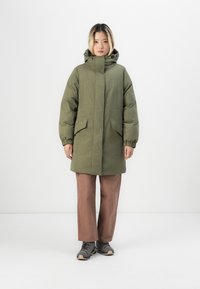 Olive green puffer coat with a high collar, elastic cuffs, and two front pockets, worn over brown wide-leg pants and gray shoes.