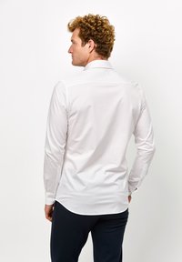 Weißes Langarmshirt mit spitzem Kragen, glatter Textur und geknöpften Manschetten; sitzt eng an der Taille und hat einen abgerundeten Saum.