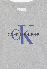 Jasnoszara tkanina z dużymi granatowymi literami „CK” oraz napisem „Calvin Klein Jeans” wydrukowanym na środku klatki piersiowej.