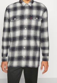 Chemise en flanelle à carreaux noir et blanc avec fermeture à boutons, deux poches poitrine et un col. Le tissu présente une texture douce et texturée.