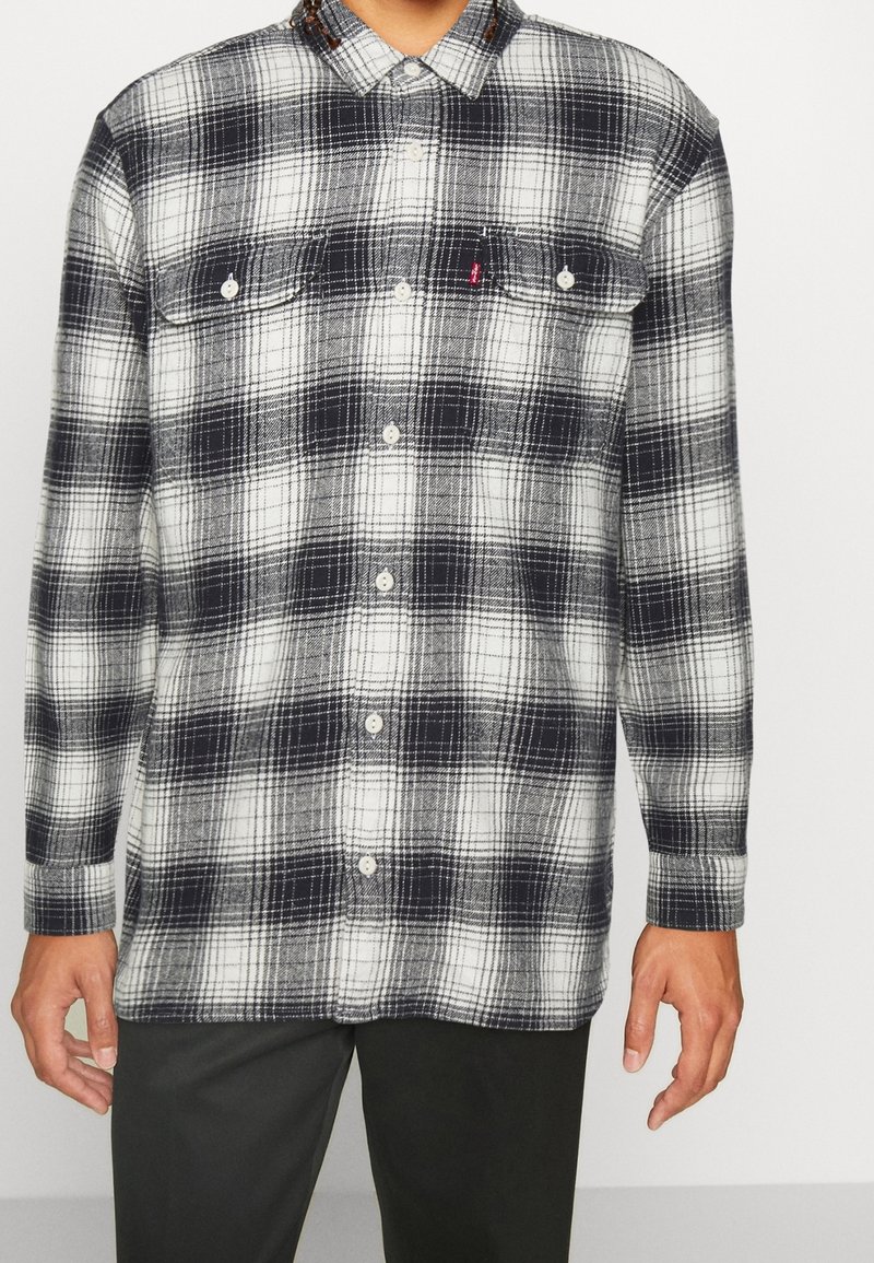 Chemise en flanelle à carreaux noir et blanc avec fermeture à boutons, deux poches poitrine et un col. Le tissu présente une texture douce et texturée.