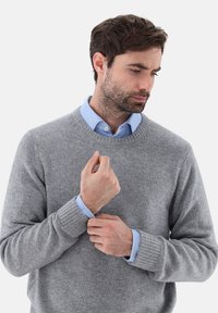 Grauer Wollpullover mit gerippten Bündchen über einem hellblauen Hemd mit Kragen. Der Mann passt seinen Hemdsärmel an und zeigt dabei die weichen Texturen.