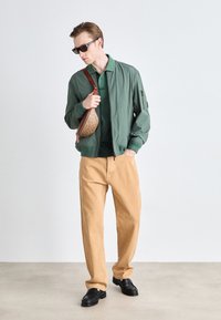 Zelená bomber bunda, tmavě zelený polo, béžové kalhoty, černé boty. Obsahuje hnědou vzorovanou crossbody tašku a sluneční brýle. Neformální outfit.