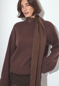 Rjav pleteni pulover z rebrastim visokim ovratnikom in dolgimi rokavi, ki ima usklajeno rjavo tkaninsko šal, zavezan okoli vratu. Teksturne površine.