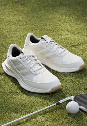 Chaussures de sport blanches sur l'herbe à côté d'un club de golf et d'une balle de golf, suggérant une utilisation pour le golf ou des activités sportives en plein air.