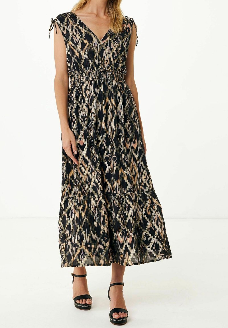 Femme portant une robe midi sans manches à col en V avec un motif abstrait noir, beige et marron, et des sandales à talons hauts noires avec bride à la cheville.