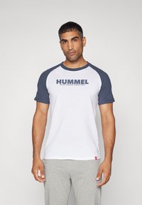 Bílý bavlněný tričko s modrými raglánovými rukávy, na kterém se nachází výrazné logo "HUMMEL" v námořnické modři přes hruď. Hladká textura, volný střih.