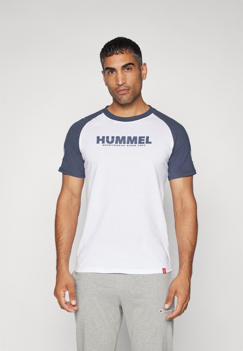 Bílý bavlněný tričko s modrými raglánovými rukávy, na kterém se nachází výrazné logo "HUMMEL" v námořnické modři přes hruď. Hladká textura, volný střih.
