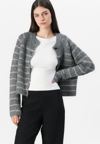 Γκρι και άσπρη ριγέ κάρντιγκαν, πλεκτή υφή, στρογγυλή λαιμόκοψη και κλείσιμο με κουμπί, φορεμένη πάνω από άσπρο τοπ και μαύρο παντελόνι.