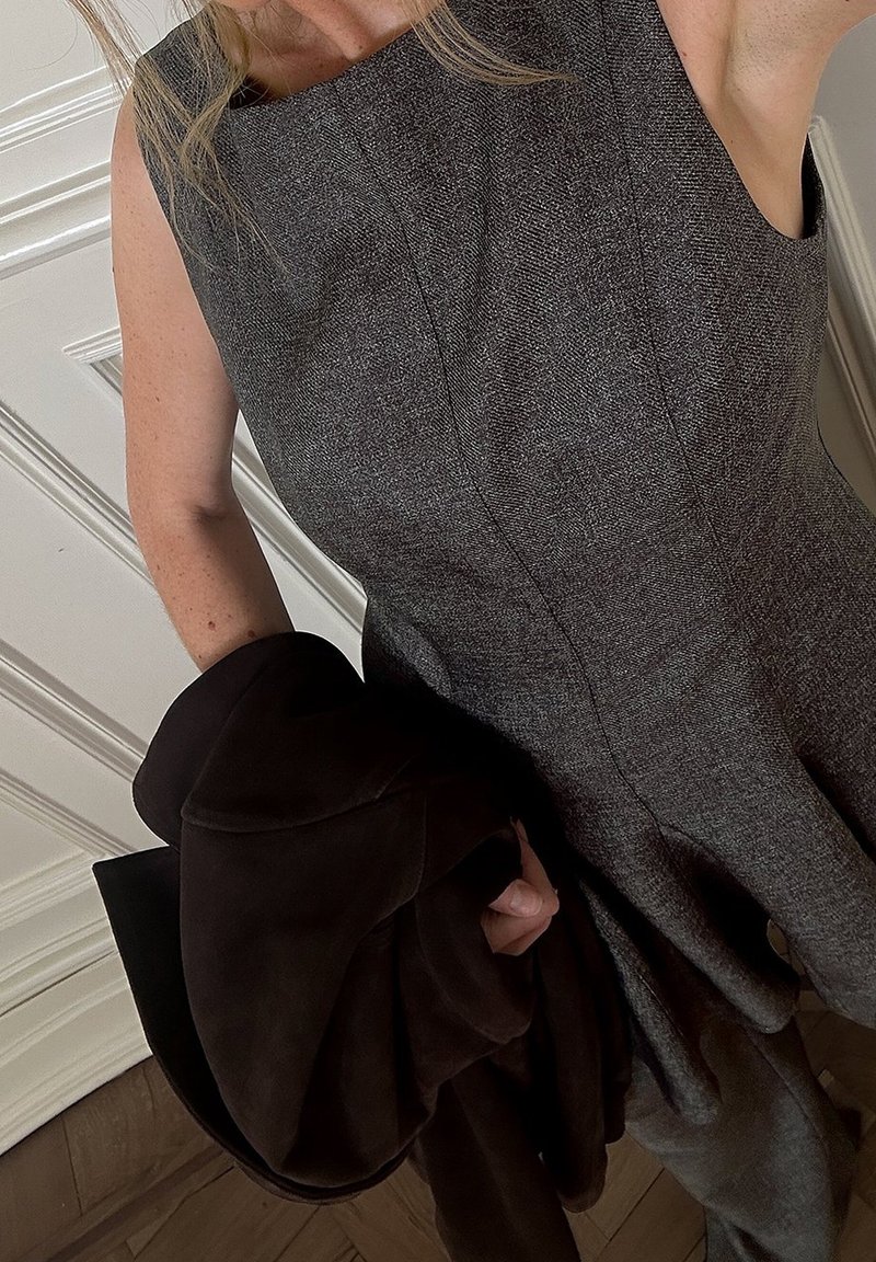 Femme portant une robe grise sans manches tenant un manteau marron foncé plié, debout près d'une porte blanche à panneaux sur un sol en bois.