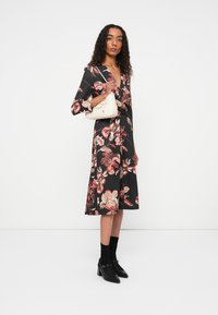 Bloemenjurk met zwarte achtergrond, voorzien van rode en beige patronen, middellange mouwen en een getailleerde taille. Geaccentueerd met een witte handtas.