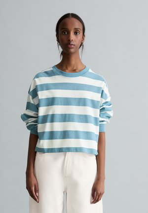 Jonge vrouw staand tegen een grijze achtergrond, gekleed in een losse blauw-wit gestreepte lange mouwen shirt en een witte broek.