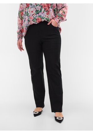 Femme portant un pantalon noir à jambes droites, un chemisier à manches longues à motif floral, et des ballerines noires pointues avec des boucles décoratives.