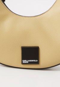 Beige leather handbag with a rounded shape, featuring a black square logo plate embossed with "KARL LAGERFELD JEANS." Smooth texture.