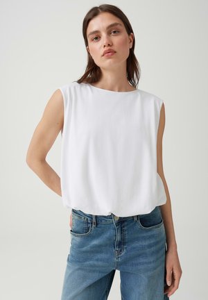 Junge Frau mit glatten braunen Haaren trägt ein ärmelloses weißes Oberteil und blaue Jeans, steht vor einem schlichten hellen Hintergrund.
