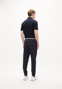 Ung mand står med ryggen til, iført en sort kortærmet polo, sorte joggers med hvide striber og hvide sneakers på en hvid baggrund.